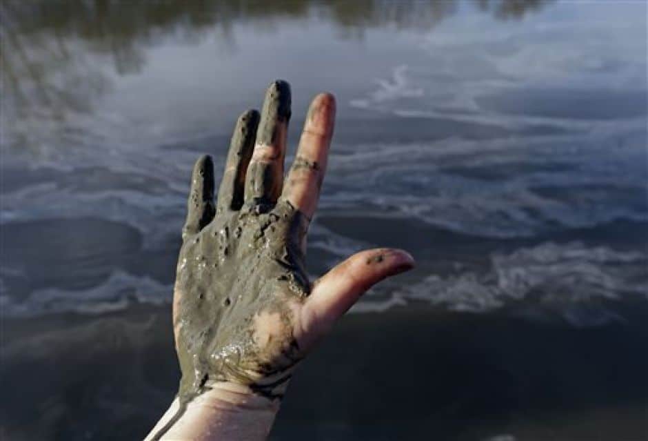 A woman's hand covered with coal ash is centered in the photo above water. The coal ash is a sludgy brownish-gray.