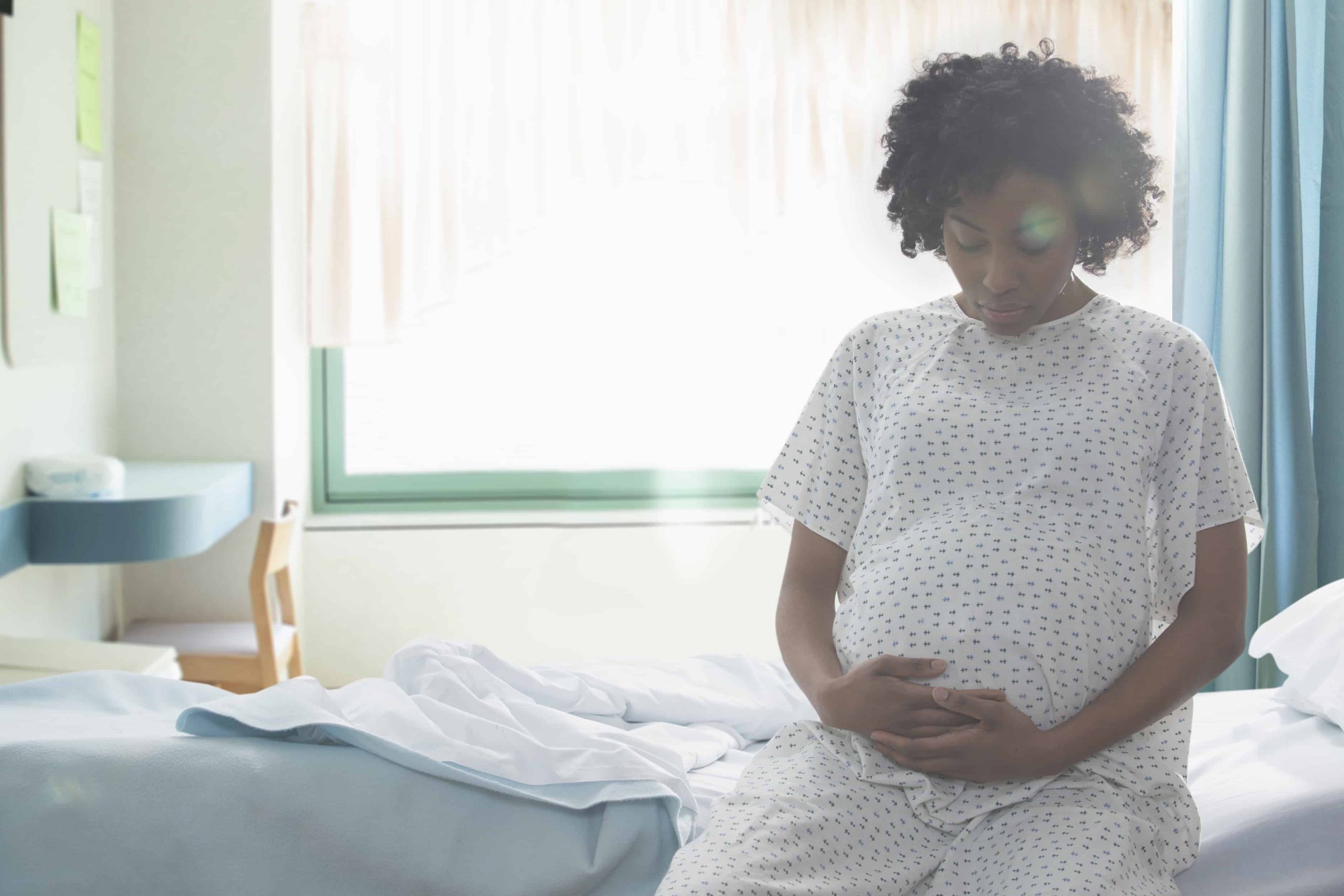 A Black pregnant woman holds her belly as she sits on a bed in a hospital.