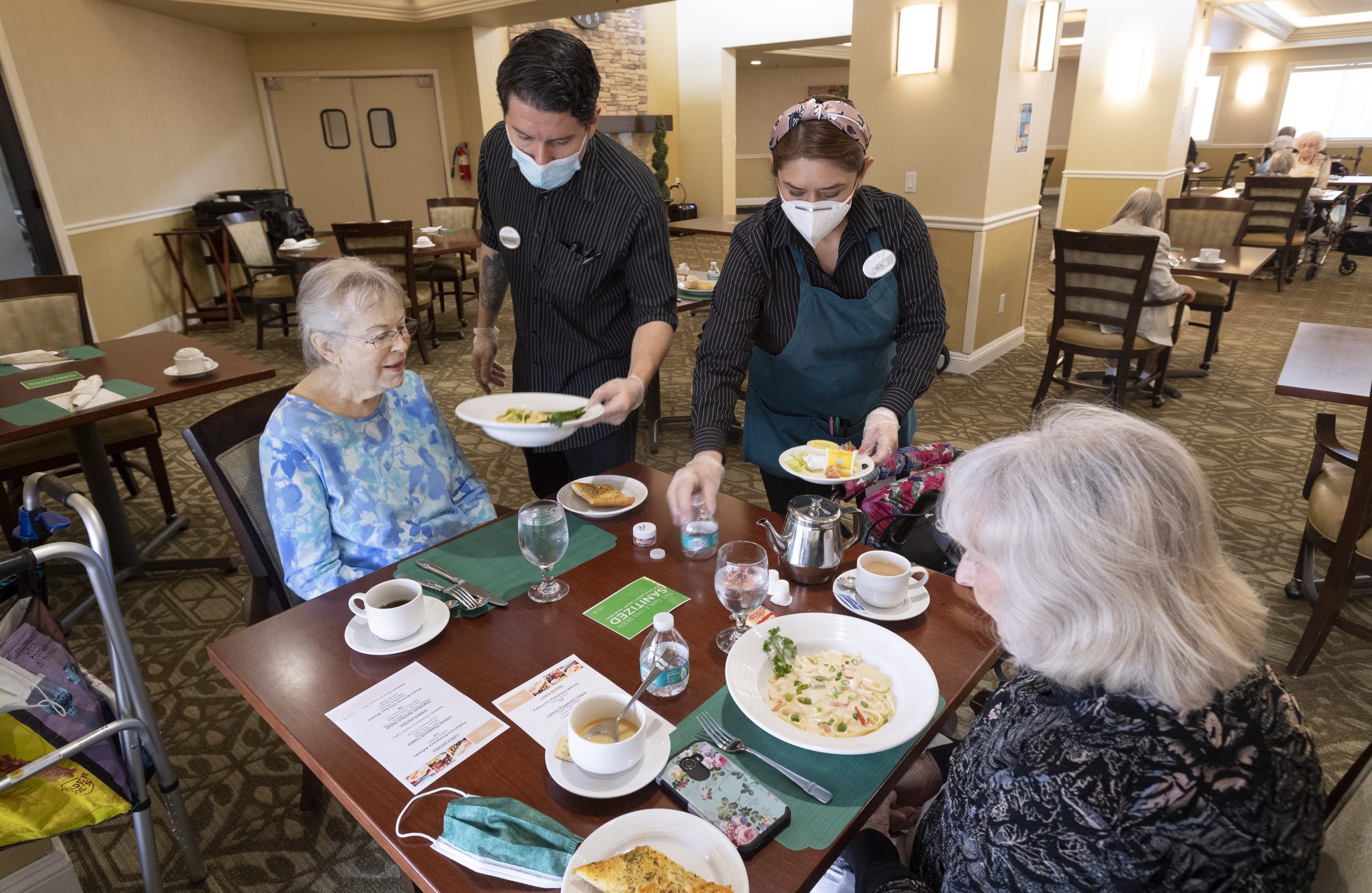 Two nursing home workers distribute pasta to two residents in the dining area of a nursing home.