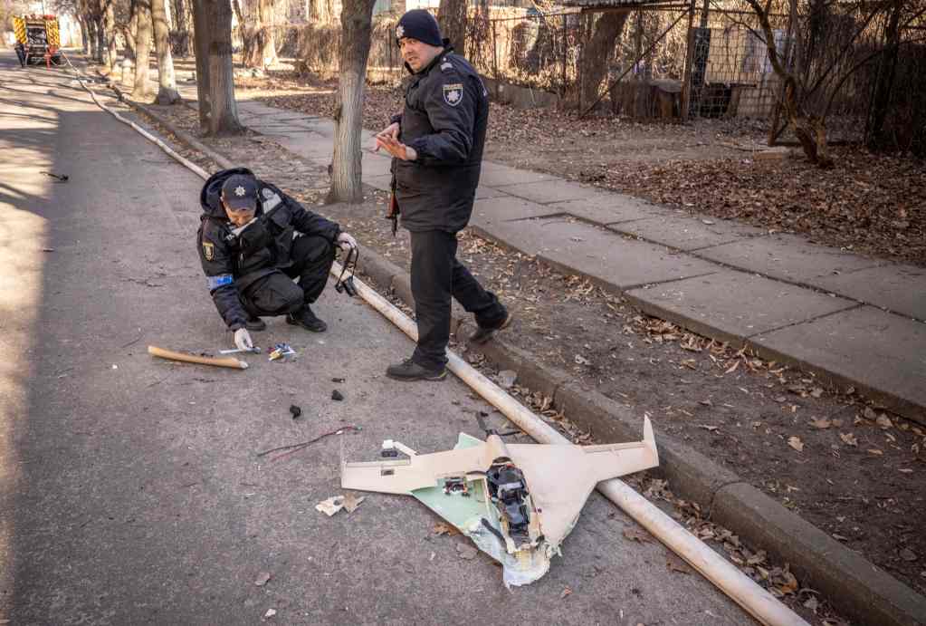 Two Ukrainian police officers examine a broken Russian drone. One officer is crouched on the ground looking at pieces of the device. The other stands near him.