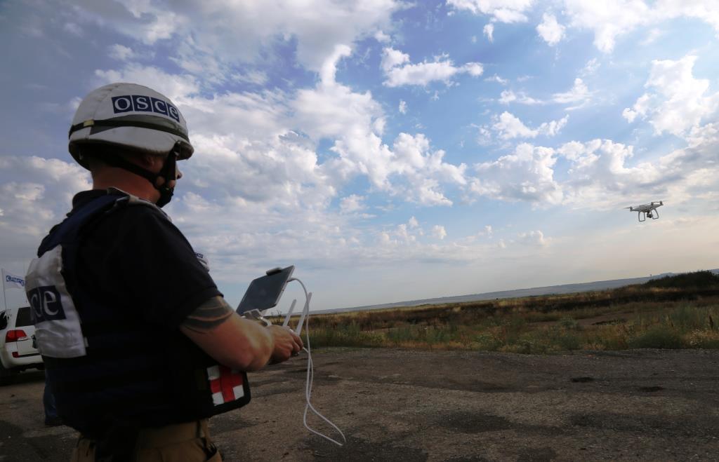 Man wearing a white OSCE helmet holds a monitoring devices to control a drone flying over a field in Ukraine.