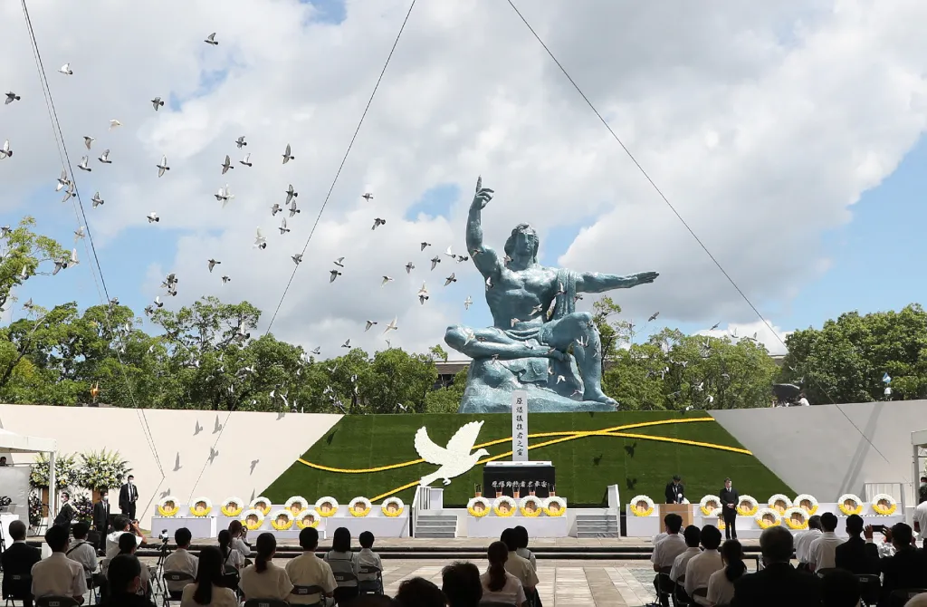 Doves flight in a blue sky with puffy clouds above a memorial services with a large blue sculpture of a man pointing to the sky with his right hand and reaching out with his left hand. The statue has closed eyes and is seated with his right leg folded under his left leg.hite and yellow wreaths a placed across the base as people look on.