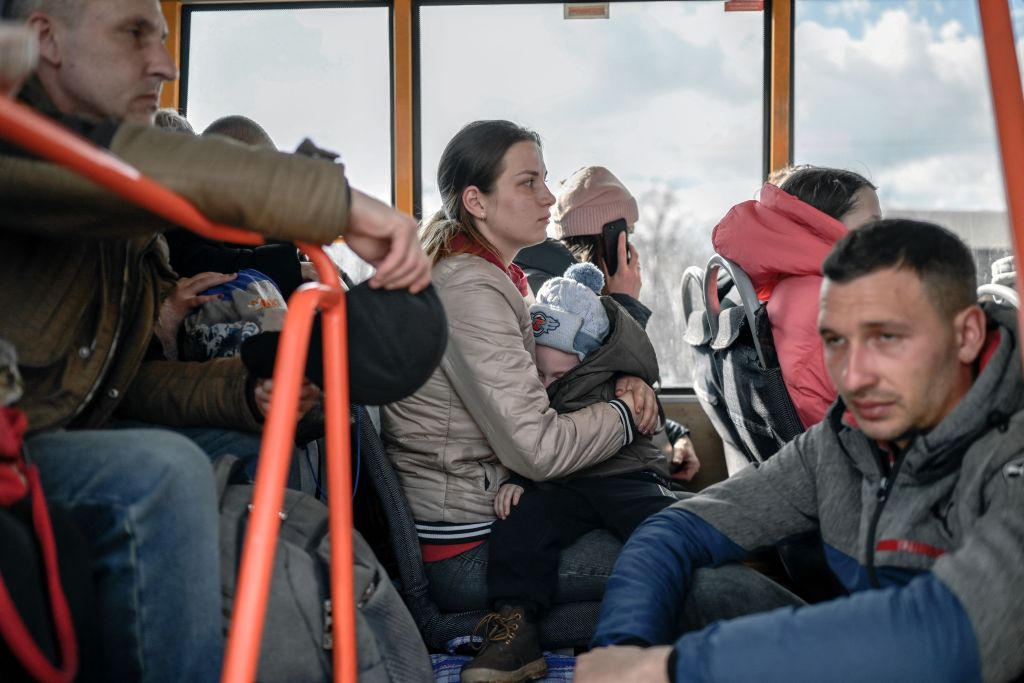 People are on a bus evacuating. A woman in a tan jacket holds a sleeping child on her lap. A man in a blue and gray jacket stares off into the distance. Several other people are crowded around.