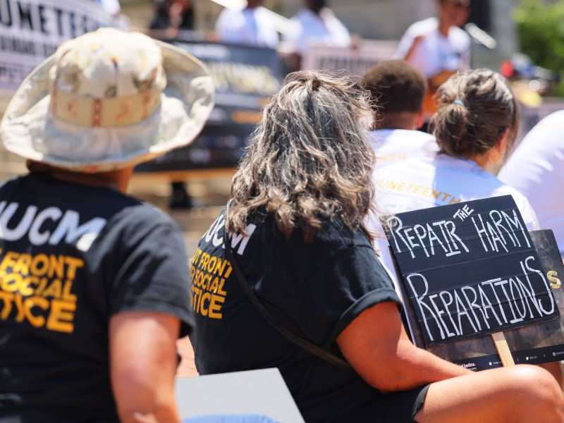 Protesters sit while holding a sign that says "Repair the harm. Reparations."