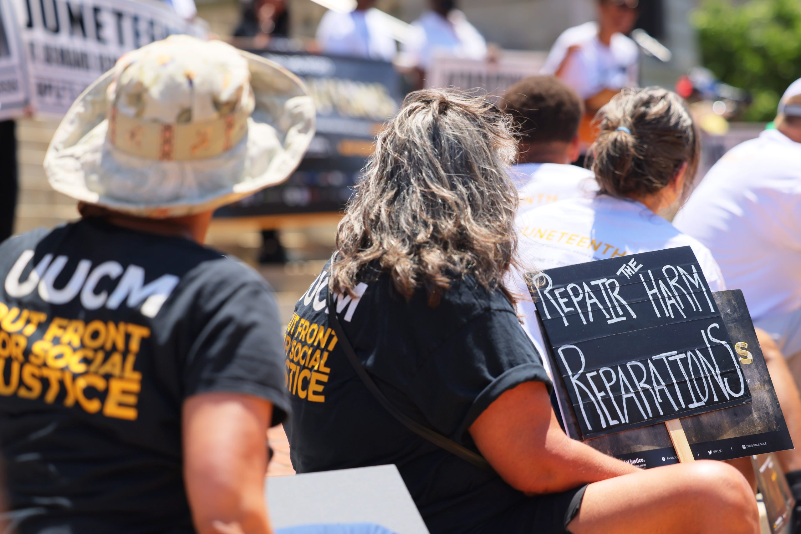 Protesters sit while holding a sign that says "Repair the harm. Reparations."