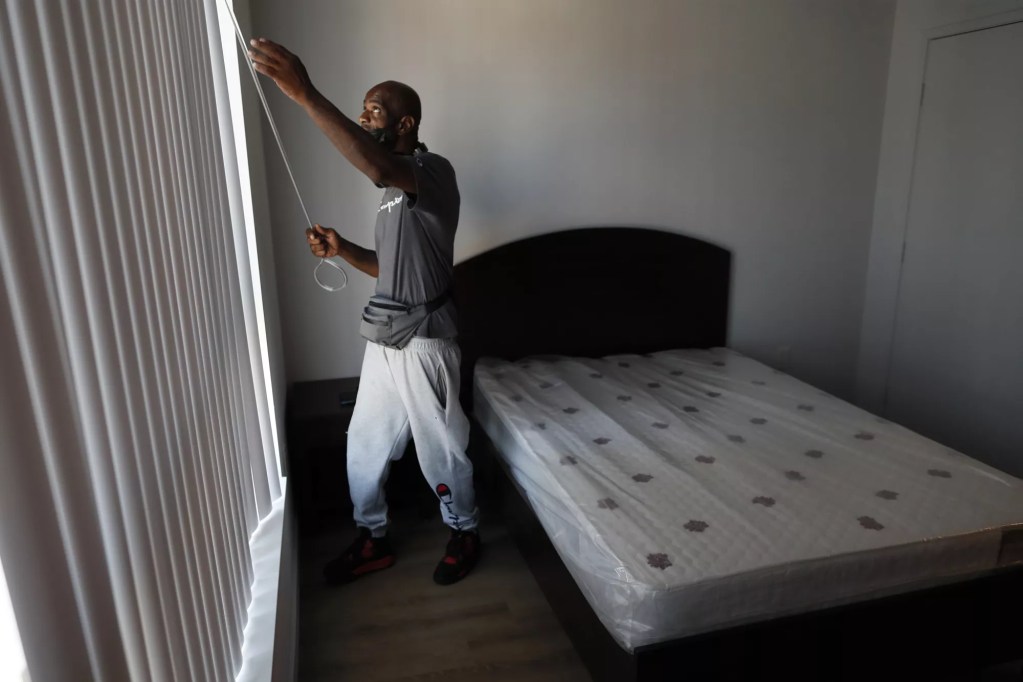 Man wearing gray shirt and white pants reaches up to close blinds. Behind him is a bed with a brown headboard and no linens.