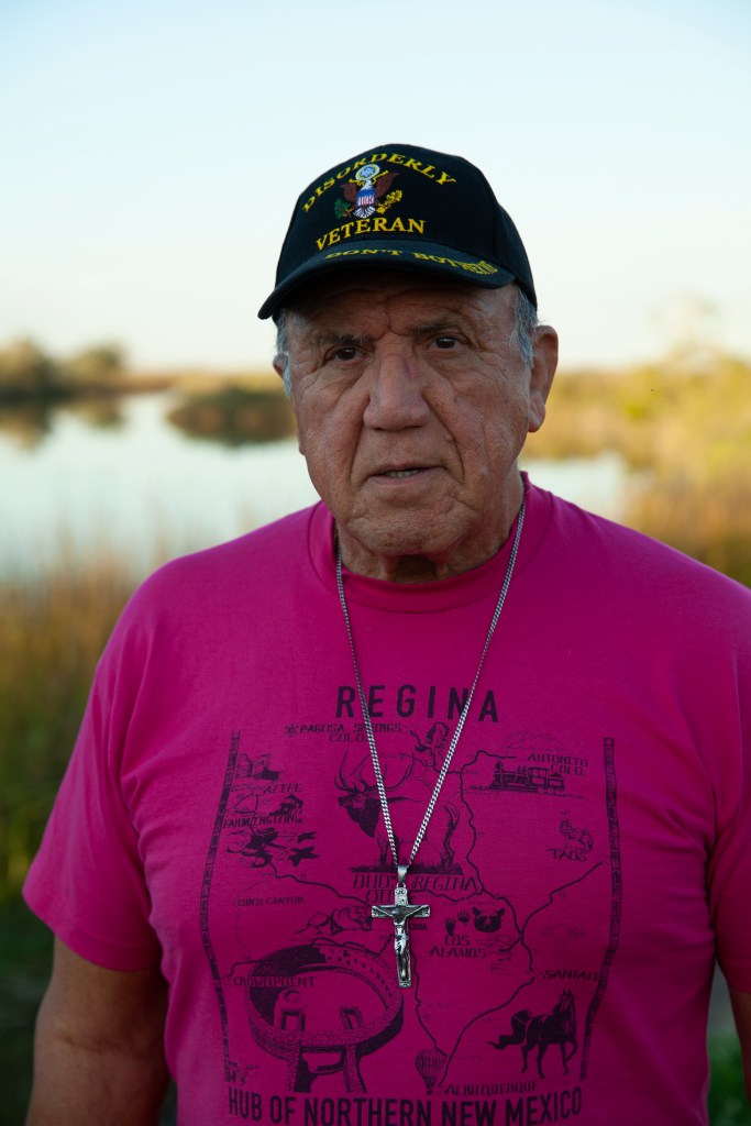 Albert White Buffalo Naquin is seen in a pink shirt with a silver cross around his neck. He is also wearing a veteran's baseball cap. 