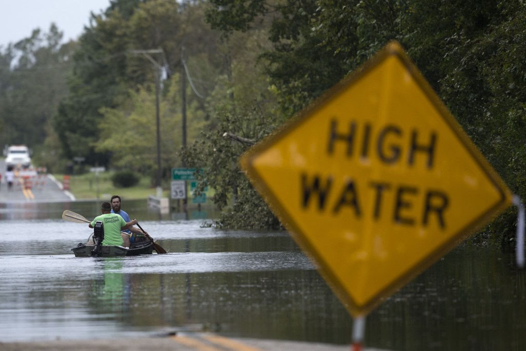 Two men in a riverboat paddle through a flooded street. A sign reading "High water" is partially submerged in the flooded street.