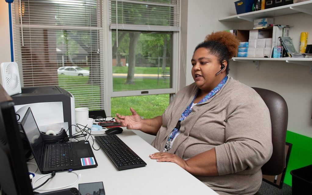 A woman sits in front of a lab top while she talks with a headset. She wears a tan cardigan with a blue and white shirt.