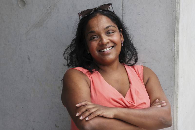 Woman with dark shoulder-length hair and sunglasses on her head smiles. Her arms are folded and she wears a coral top.