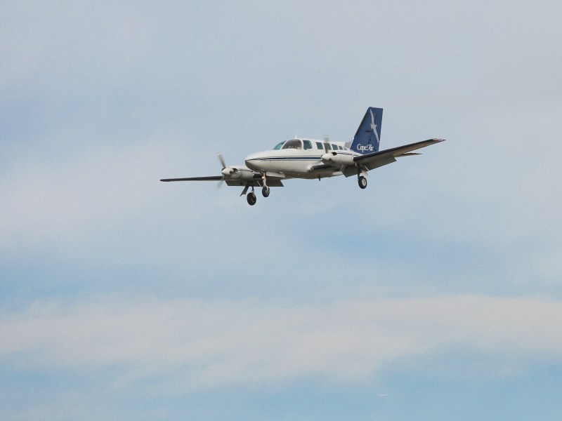 A piston-engine aircraft flies against a cloudy sky