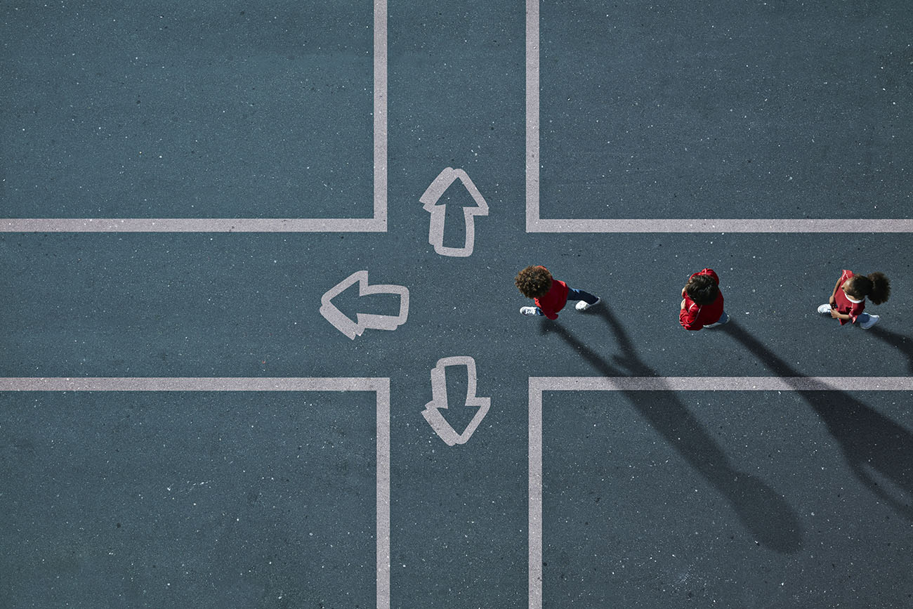 Group of children photographed from above with arrows pointing in different directions painted on the tarmac in which they walk.