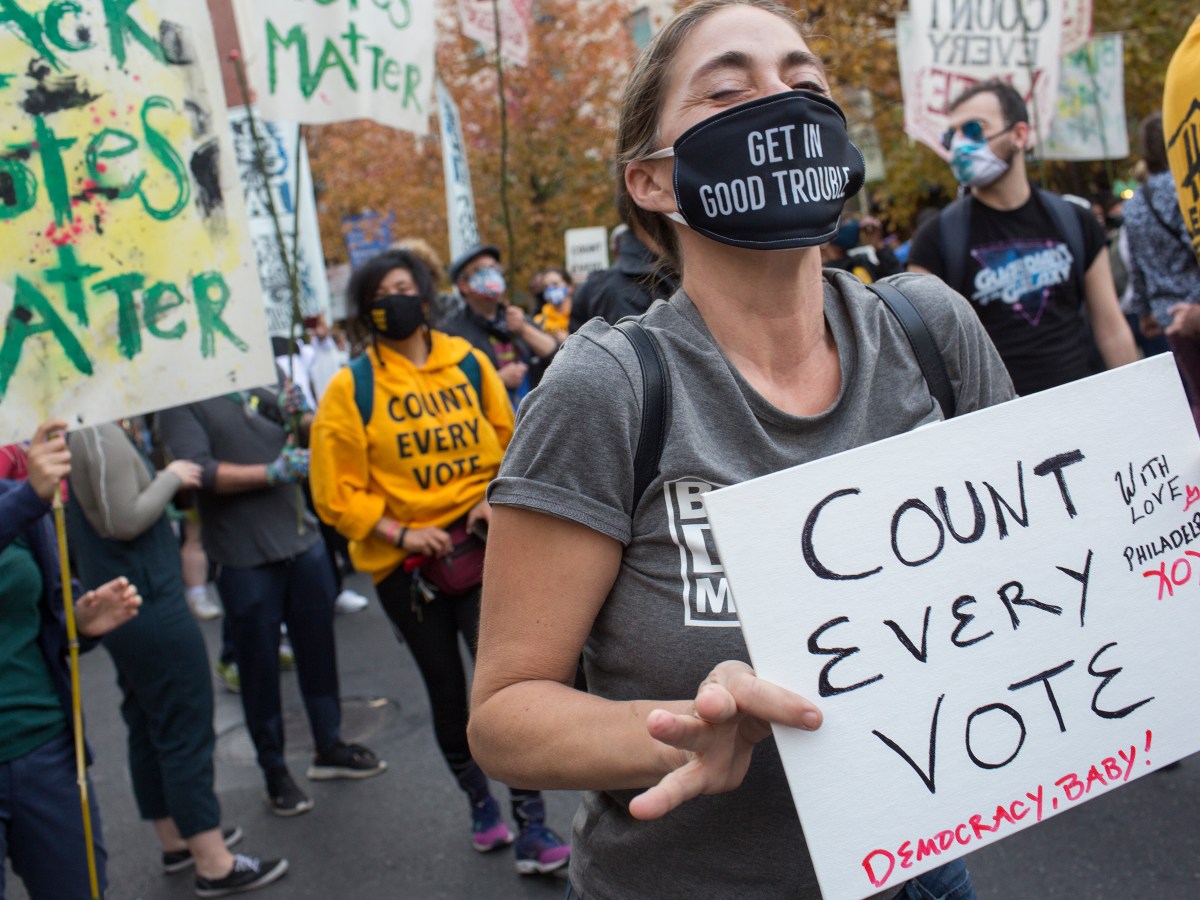 A woman standing in front of other protesters holding signs wears a gray shirt and a black face mask that says "Get in Good Trouble" on it and holds a white homemade cardboard sign that says "Count Every Vote" in black letters, with the words, "Democracy, Baby!" and "With love, Philadelphia XOXO" in smaller read letters.