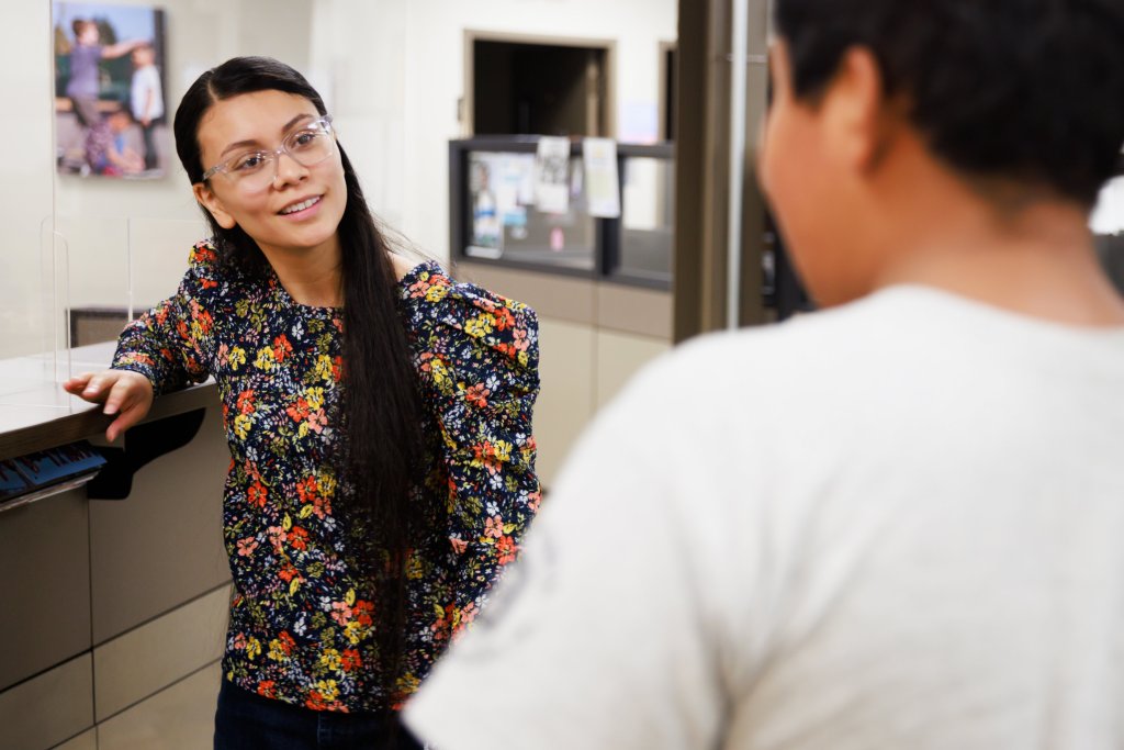Jessica Llamas smiles at the student she's helping, who is visible from behind. Llamas is wearing a flowered shirt and glasses, and she has long dark hair.