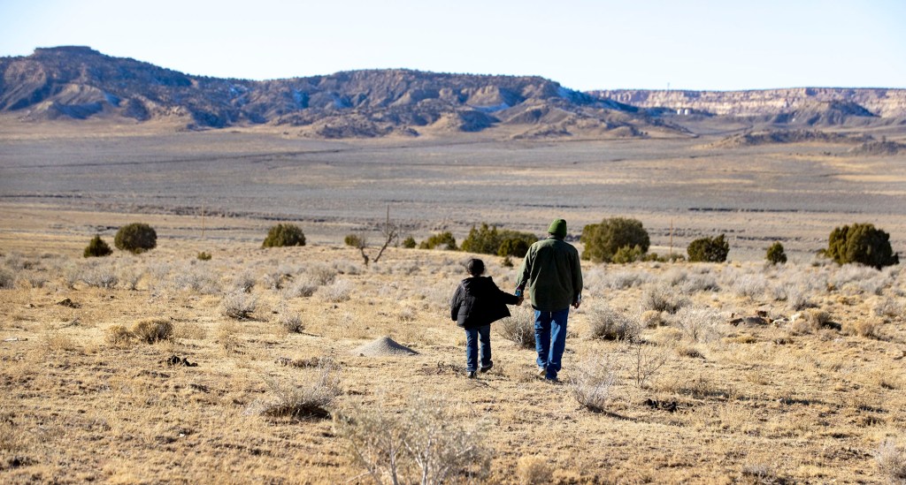 Earl Tulley and grandson Naabaahii hold hands as they walk away from the camera, toward hills.