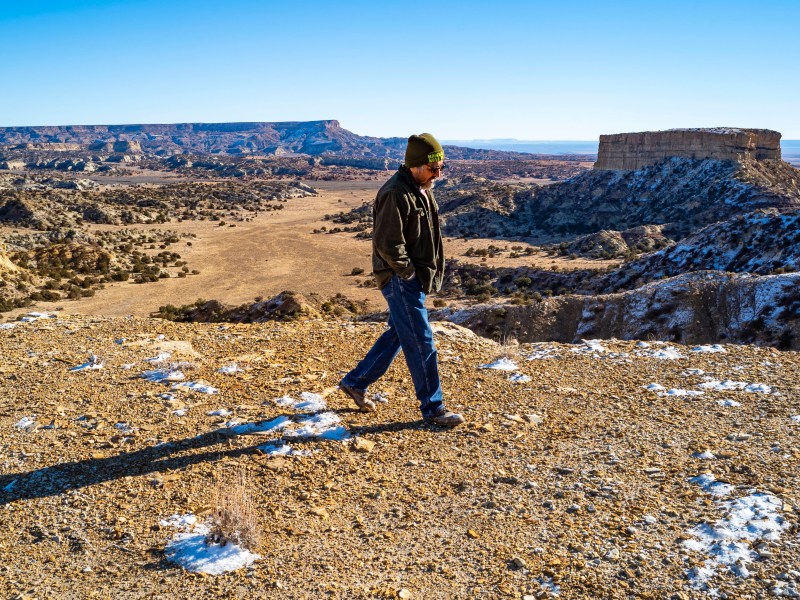 Earl Tulley walks contemplatively, looking down, against a backdrop of mesas with a dusting of snow.