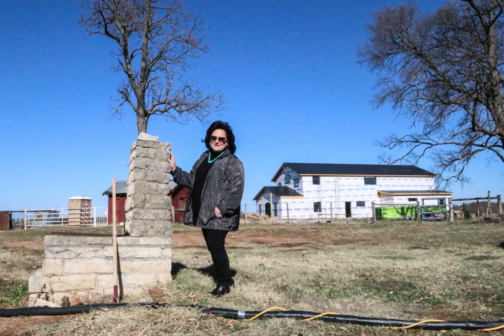 Teresa Rutherford stands on her property with her under-construction home in the background, a rural setting with two trees. 