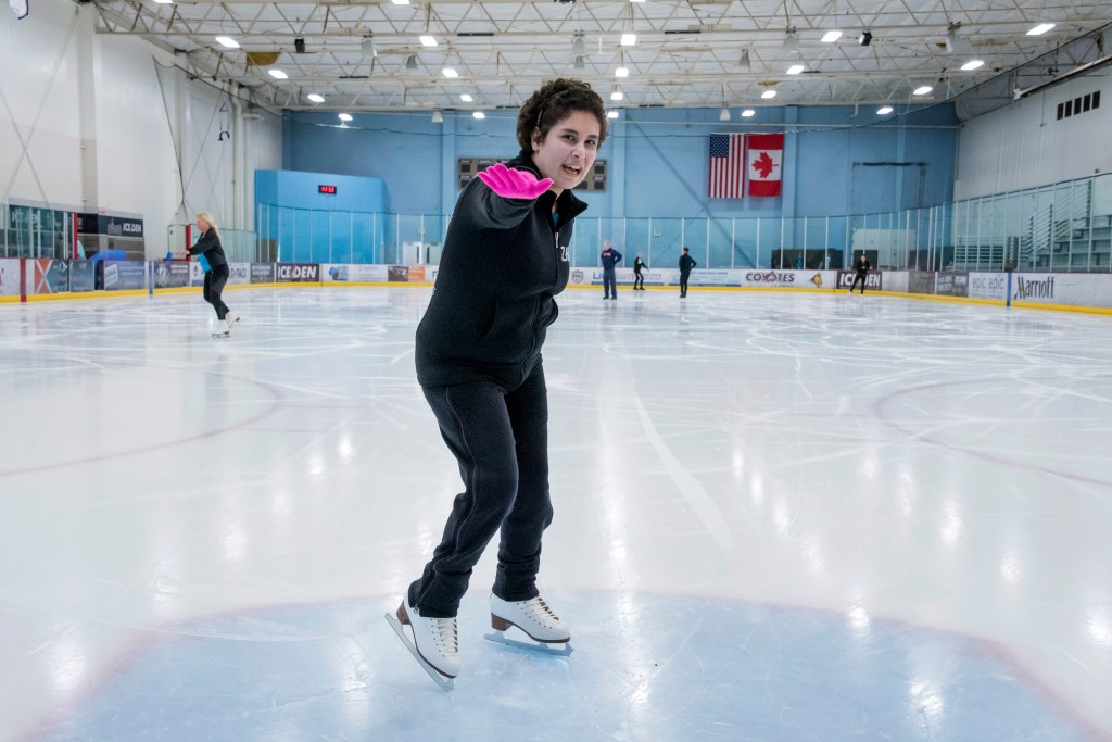 Zainab Edwards points her right arm forward while standing on the ice. She is wearing black pants, a black sweatshirt and pink gloves.