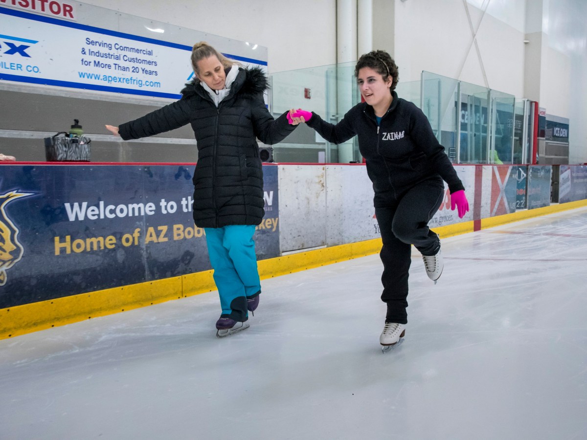 Zainab Edwards holds hands with her teacher Tammy Jimenez as they skate. Zainab wears a black sweatshirt with her name on the upper left. Her left knee is bent. Her instructor wears blue pants and a black coat. She is watching Zainab as they skate.