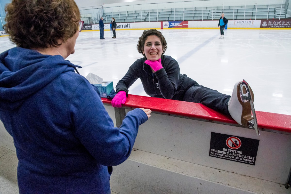 Zainab Edwards puts her left leg on the edge of the ice rink to stretch as her mom talks with her. Zainab has her hand under her chin and is smiling.