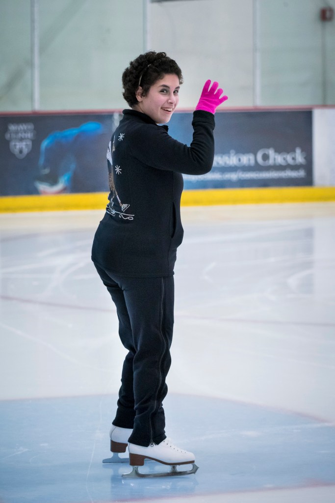 Zainab Edwards waves while standing on the ice. She is wearing black pants, a black sweatshirt and pink gloves. She has short dark hair and is smiling.