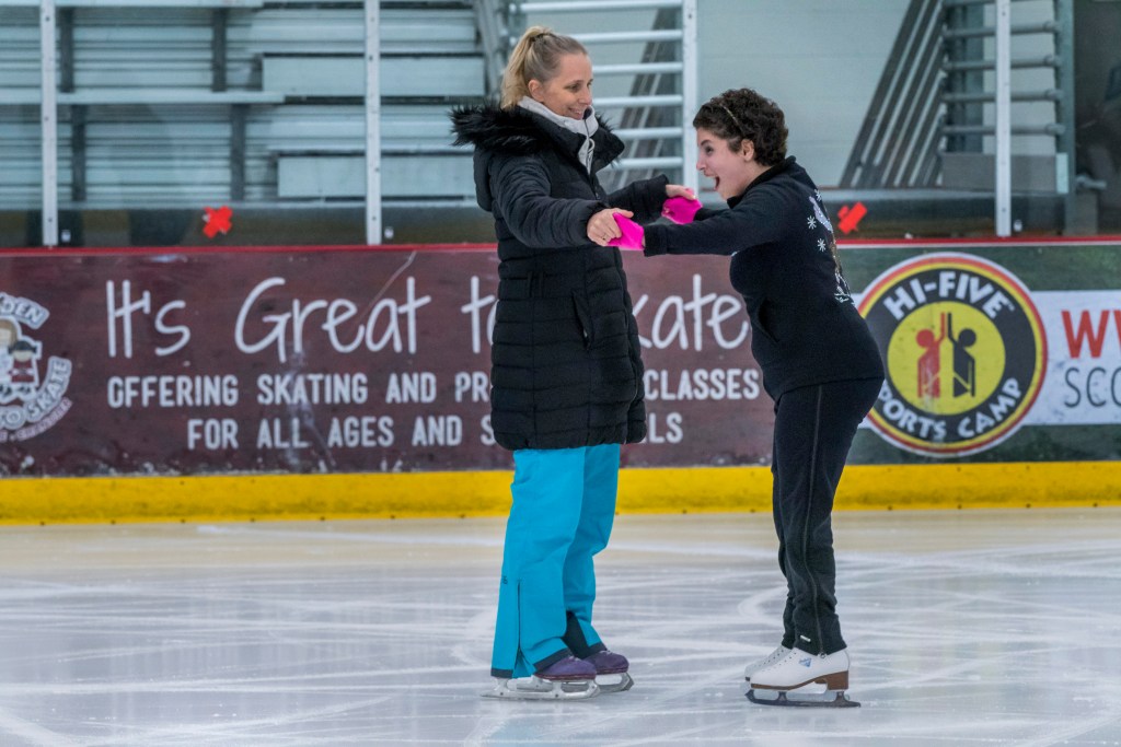 Zainab Edwards skates while holding both hands with her teacher Tammy Jimenez. Zainab has excited look on her face. She wears black pants and a black sweatshirt. Her teacher wears blue pants and a black coat.