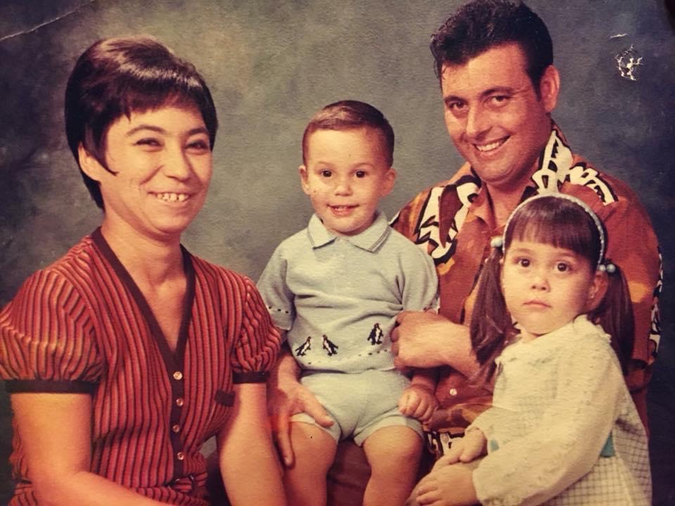 A family portrait of the Carson family from 1971. Joaquin's mother is on the left. She has short dark hair and is wearing a short-sleeved shirt with red and black strips. Joaquin is in the middle. He is smiling and wearing a light blue shirt with penguins along the bottom. On the right is Joaquin's father. He is wearing a brown and white patterned shirt. He has short dark hair and is smiling. He is holding Joaquin's left arm. In front of Joaquin's dad is Diana. She has her dark hair in pigtails and wears a headband. She is wearing a white top and is not smiling.