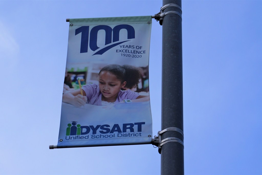 A advertisement for the Dysart Unified School District hangs from a pole. It reads '100 Years of Excellence 1920-2020'.