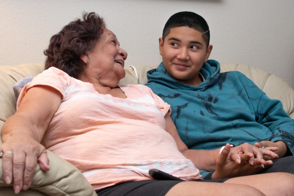 Guadalupe Hernandez, seated on a small couch beside Frances Yduarte, smiles at her as she beams at him and holds his hand.