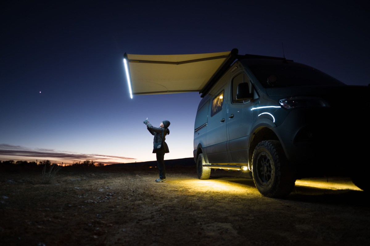 A woman stands looking at her phone next to her van, the only light comes from the van and the sky behind her.