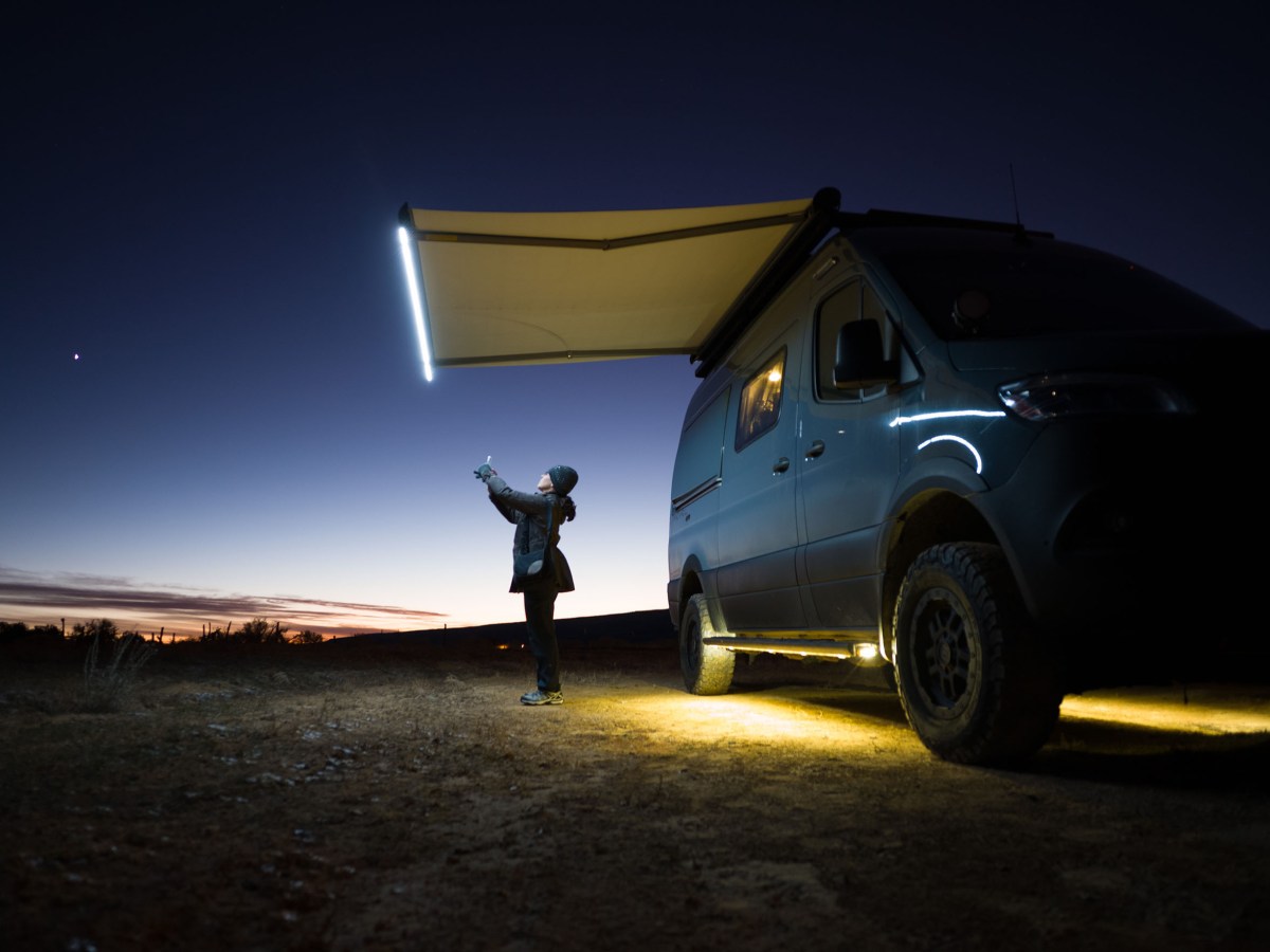 A woman stands looking at her phone next to her van, the only light comes from the van and the sky behind her.