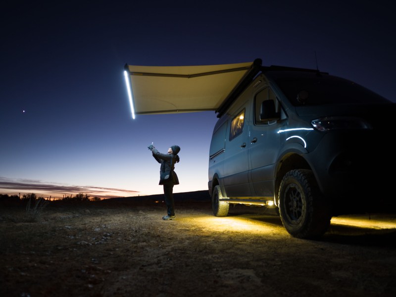 A woman stands looking at her phone next to her van, the only light comes from the van and the sky behind her.