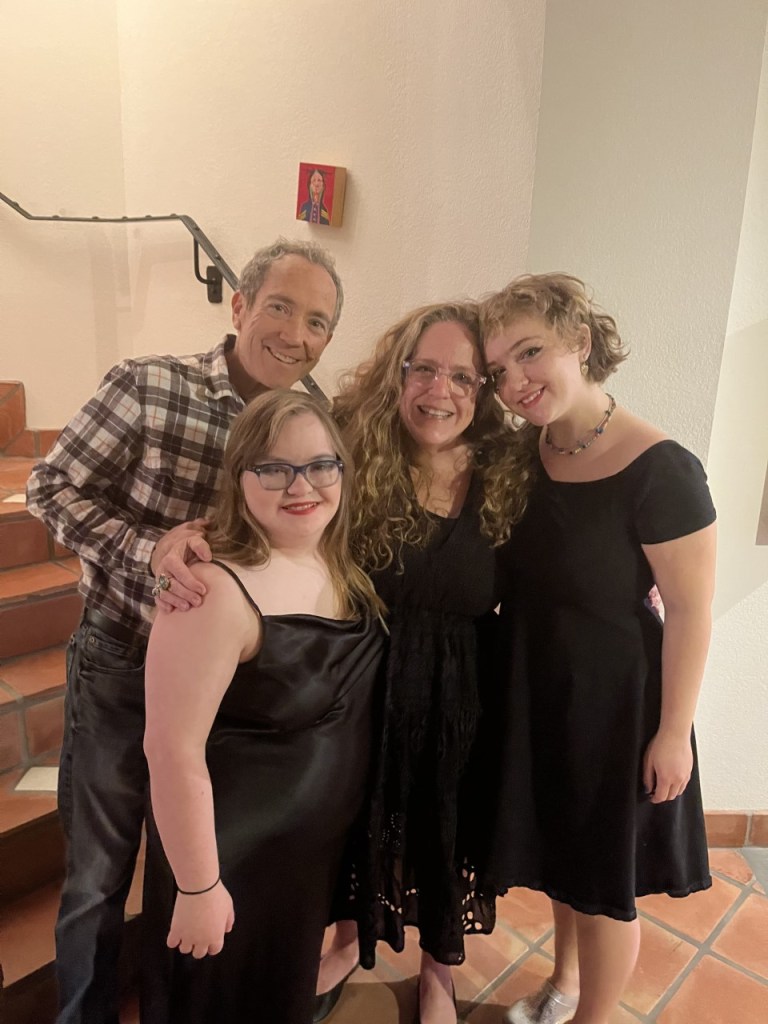 The author, Amy Silverman poses with her family at the base of a staircase. In the front is Sophie who has long hear, wears glasses and is wearing a sleeveless black dress. Amy's husband Ray Stern is behind Sophie. He wears a plaid shirt and jeans. He has his right hand on Sophie's right shoulder. Amy is in the middle. She has long wavy hair and wears glasses. She also is wearing a black dress. On the right, is Annabelle, who has her arm around Amy. She has short hair and is wearing a black dress.