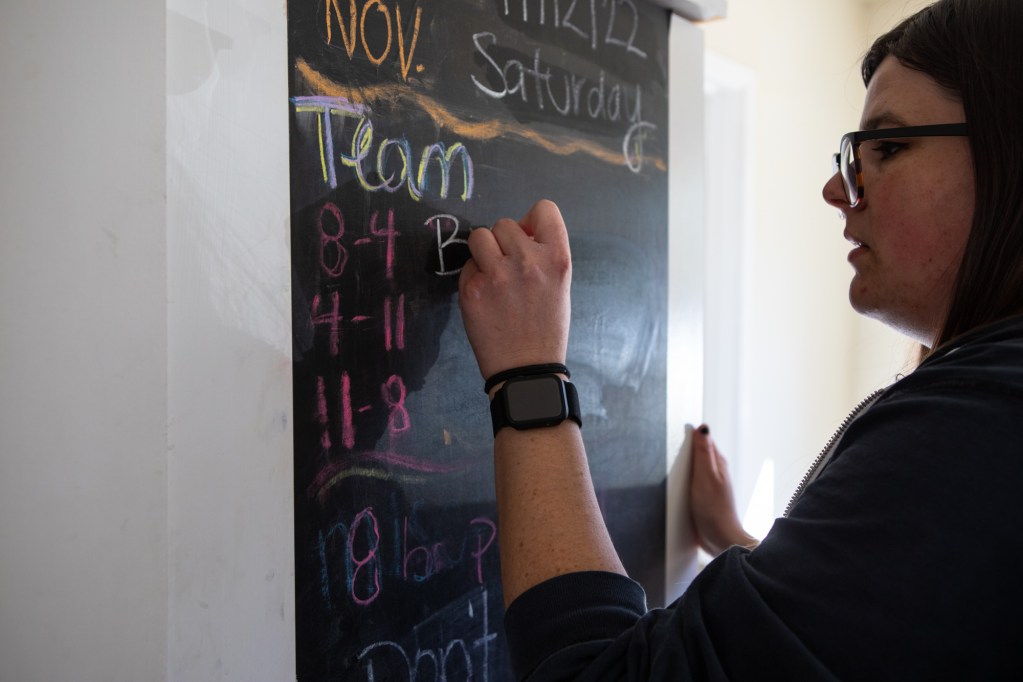 Brittany Merrill writes on a blackboard with her left hand. She is wearing a black top and glasses.