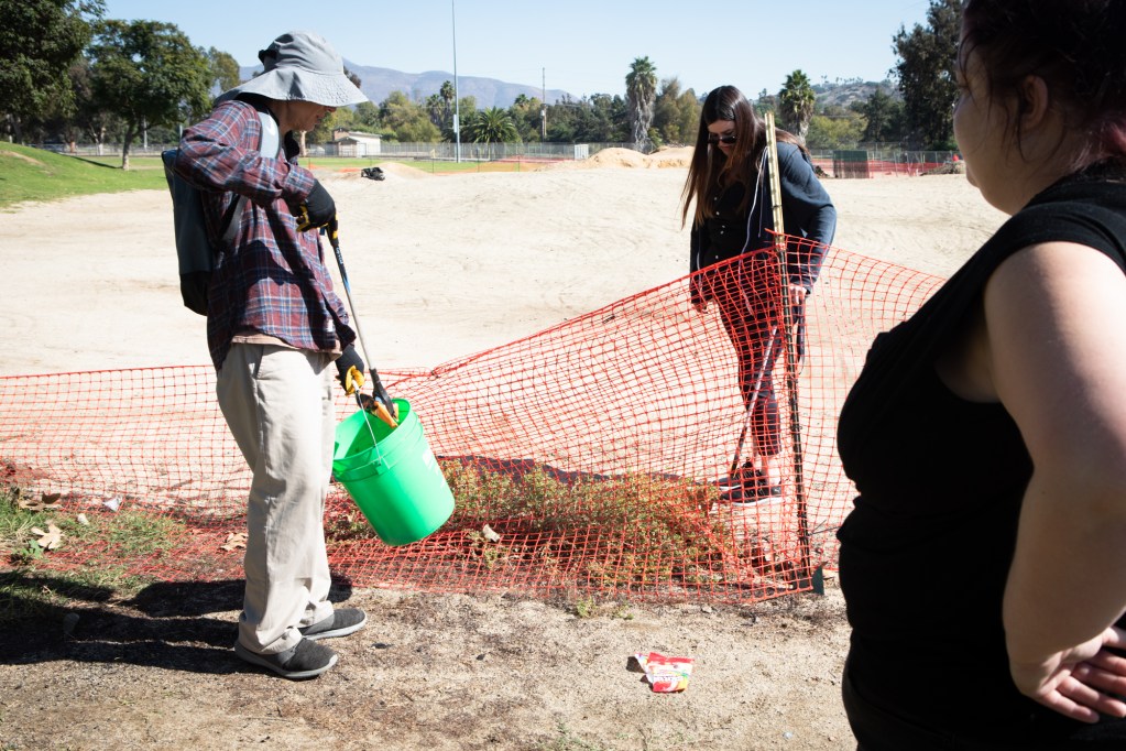 Joaquin Carson holds a green bucket and a trash grabber. He is picking up trash at a park. He wears a gray brimmed hat, a maroon and blue plaid shirt and tan pants.