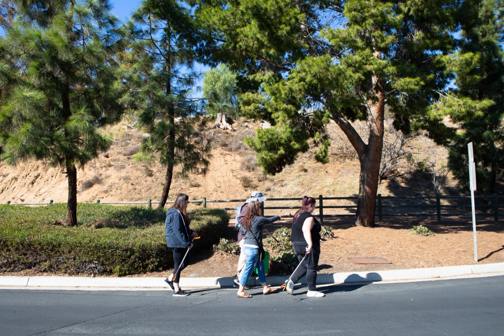 Joaquin Carson, his sister and caregivers walk along a paved road. Behind them are three pine trees, shrubs and a wooden fence. Behind the fence is a hill. His caretakers are carrying metal grabbers with orange handles.