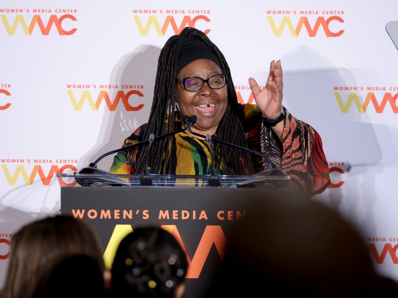 Loretta J. Ross stands behind a podium giving a speech at the Women's Media Awards. She has one arm raised and is speaking animatedly.