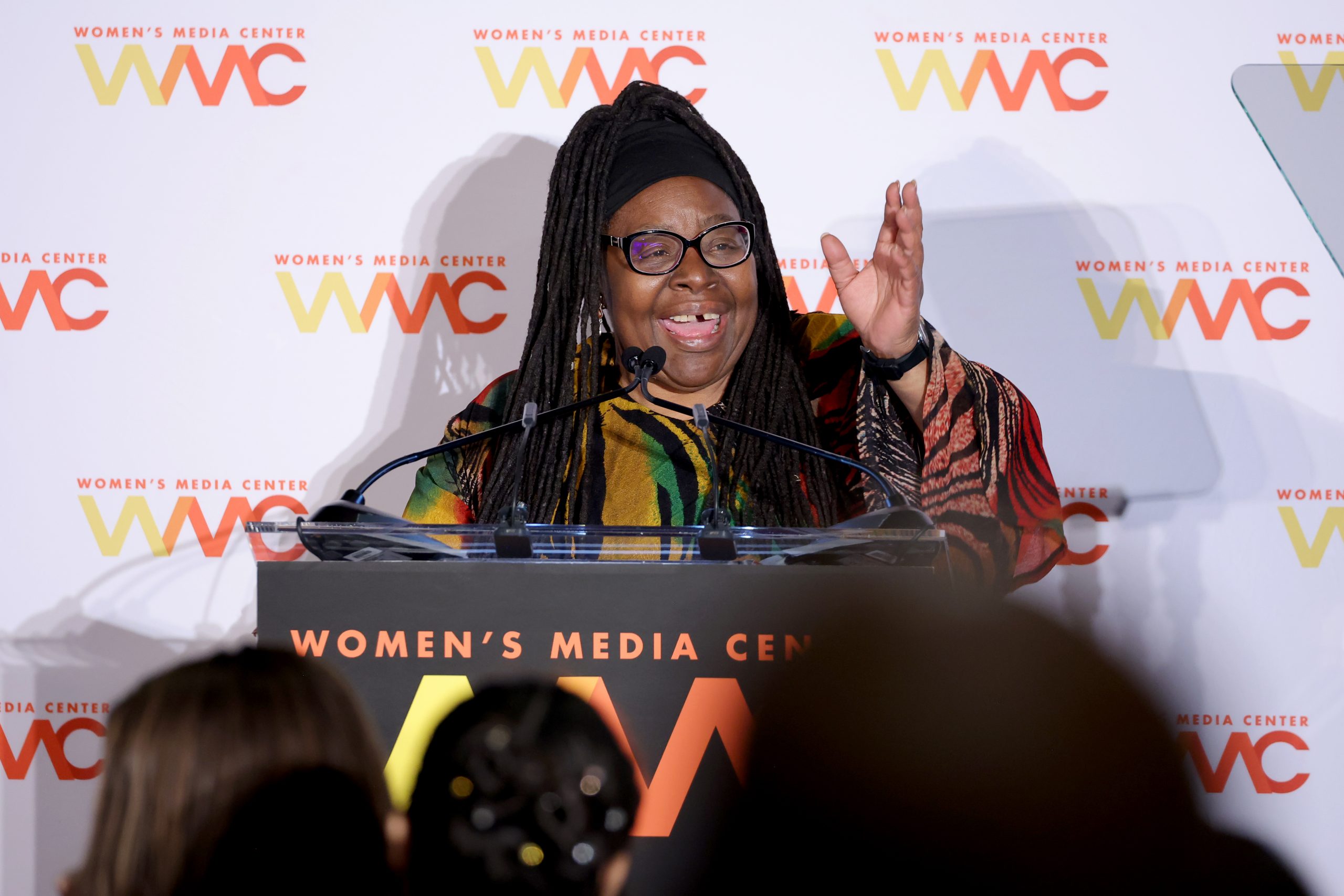 Loretta J. Ross stands behind a podium giving a speech at the Women's Media Awards. She has one arm raised and is speaking animatedly.