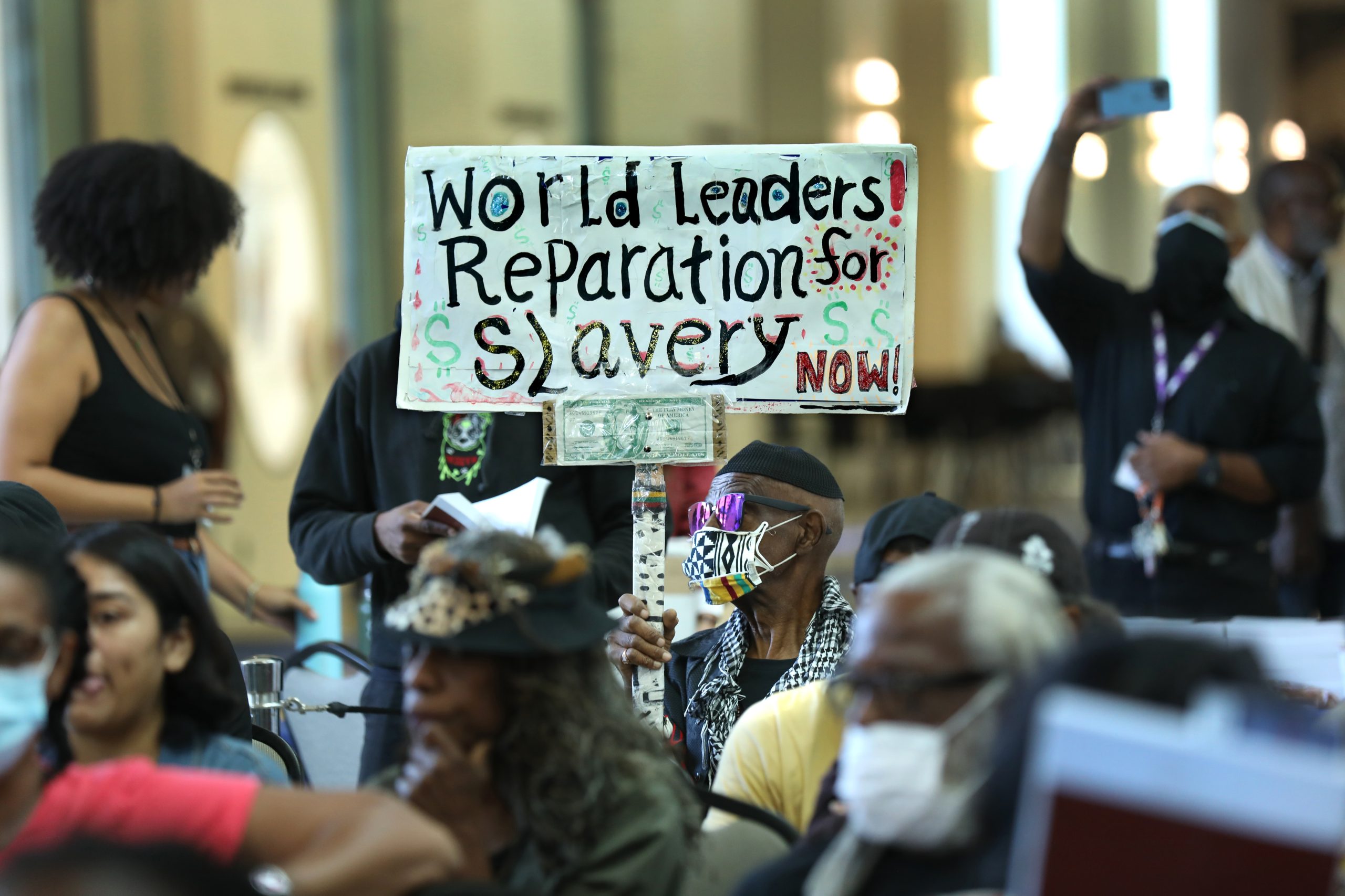 A seated man wearing a mask holds a sign that says "World leaders! Reparations for slavery now!" during a California reparations task force meeting.