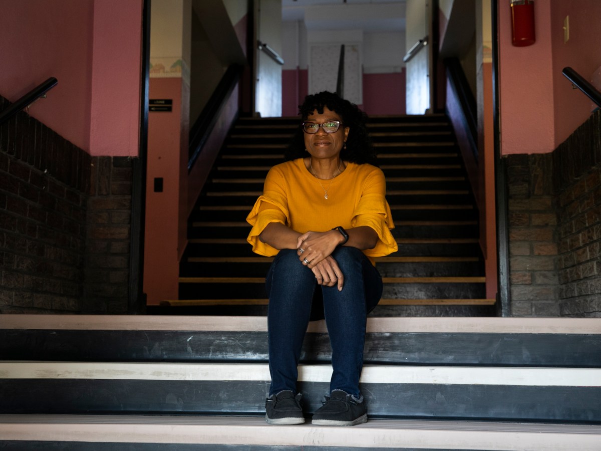 ReShonda Young, wearing a yellow-orange suit and blue pants, sits between two levels of stairs. She is wearing glasses and smiling.