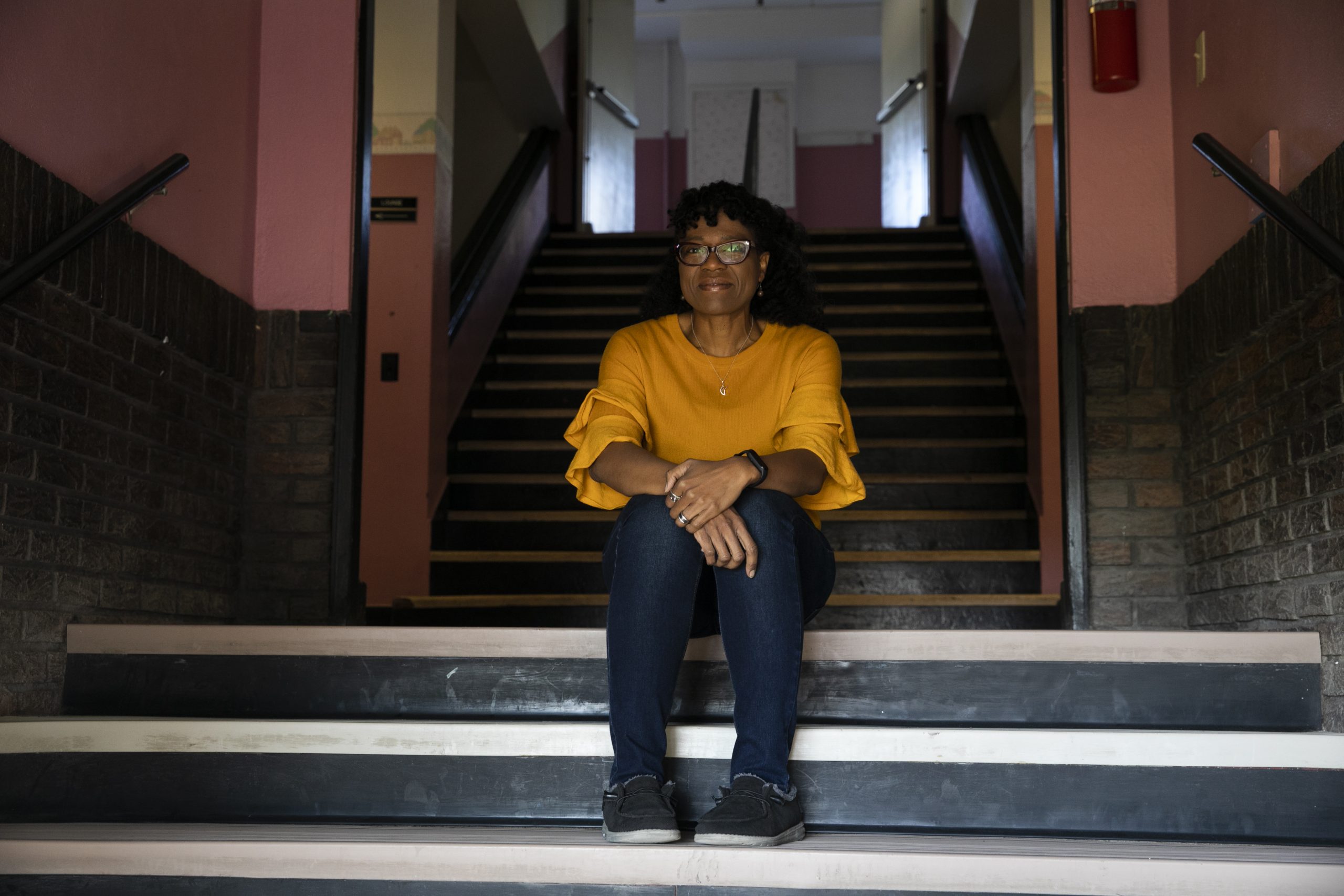 ReShonda Young, wearing a yellow-orange suit and blue pants, sits between two levels of stairs. She is wearing glasses and smiling.