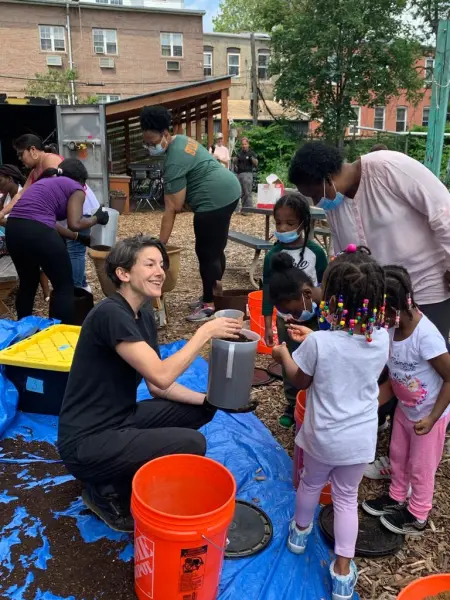 Sara Perl Egendorf is bent down as she shows a group of young children and one adult soil that has been collected in a rubber container.