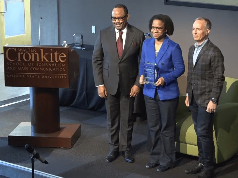 Four people stand next to a brown wooden lectern that says "Walter Cronkite School of Journalism and Mass Communication, Arizona State University," two of them holding glass awards.