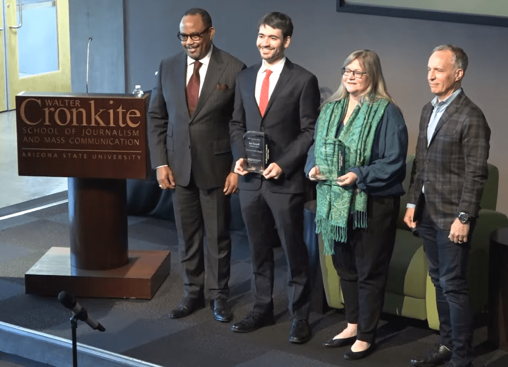 Four people stand next to a brown wooden lectern that says "Walter Cronkite School of Journalism and Mass Communication, Arizona State University," two of them holding glass awards.