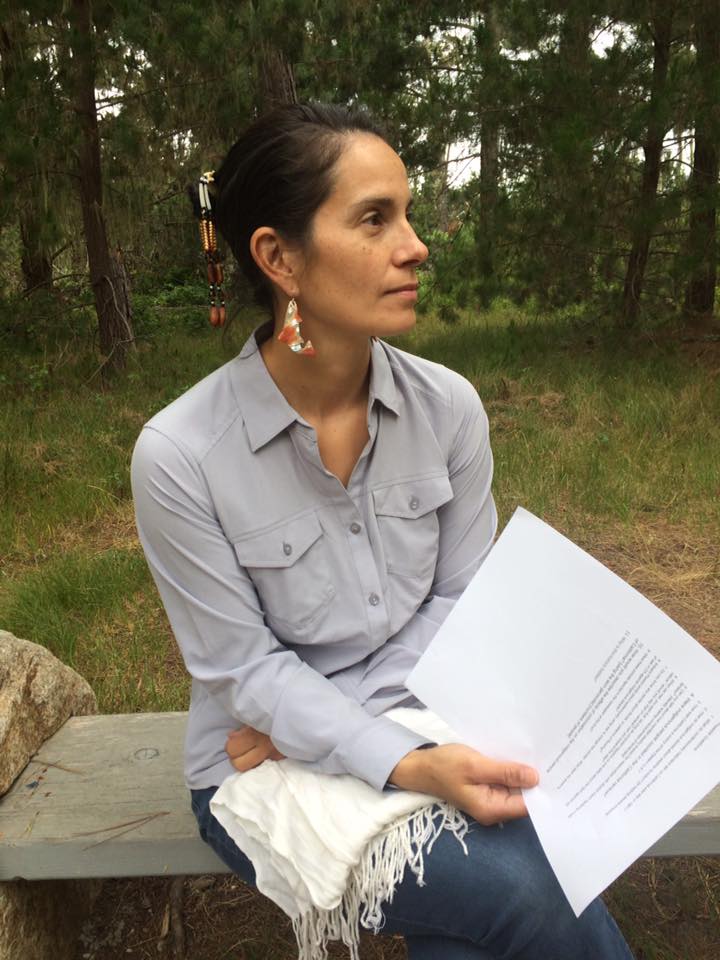 Kouslaa Kessler-Mata sits in a park on a stone bench. She is wearing a light blue button up shirt and jeans, is looking to the side, and holding a sheet of paper with several lines of text.