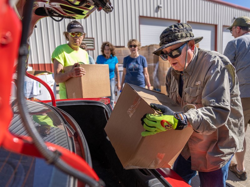 A person wearing a bucket hat, jacket, and neon green gloves places a cardboard box into the trunk of a red car. Behind them, two women wearing blue shirts and a man wearing a neon green shirt holding another box stand waiting.