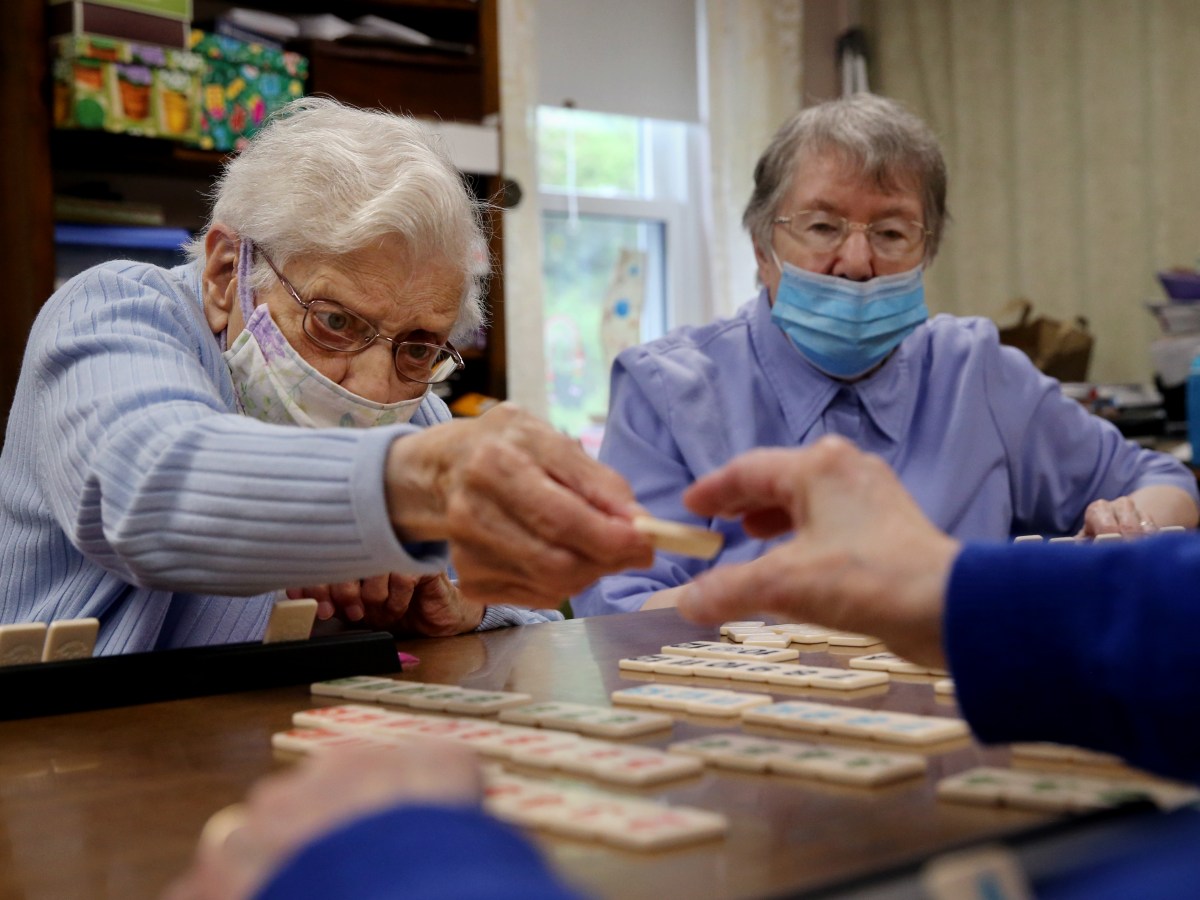 two nursing home residents play a board game at a table. they both wear blue paper face masks.
