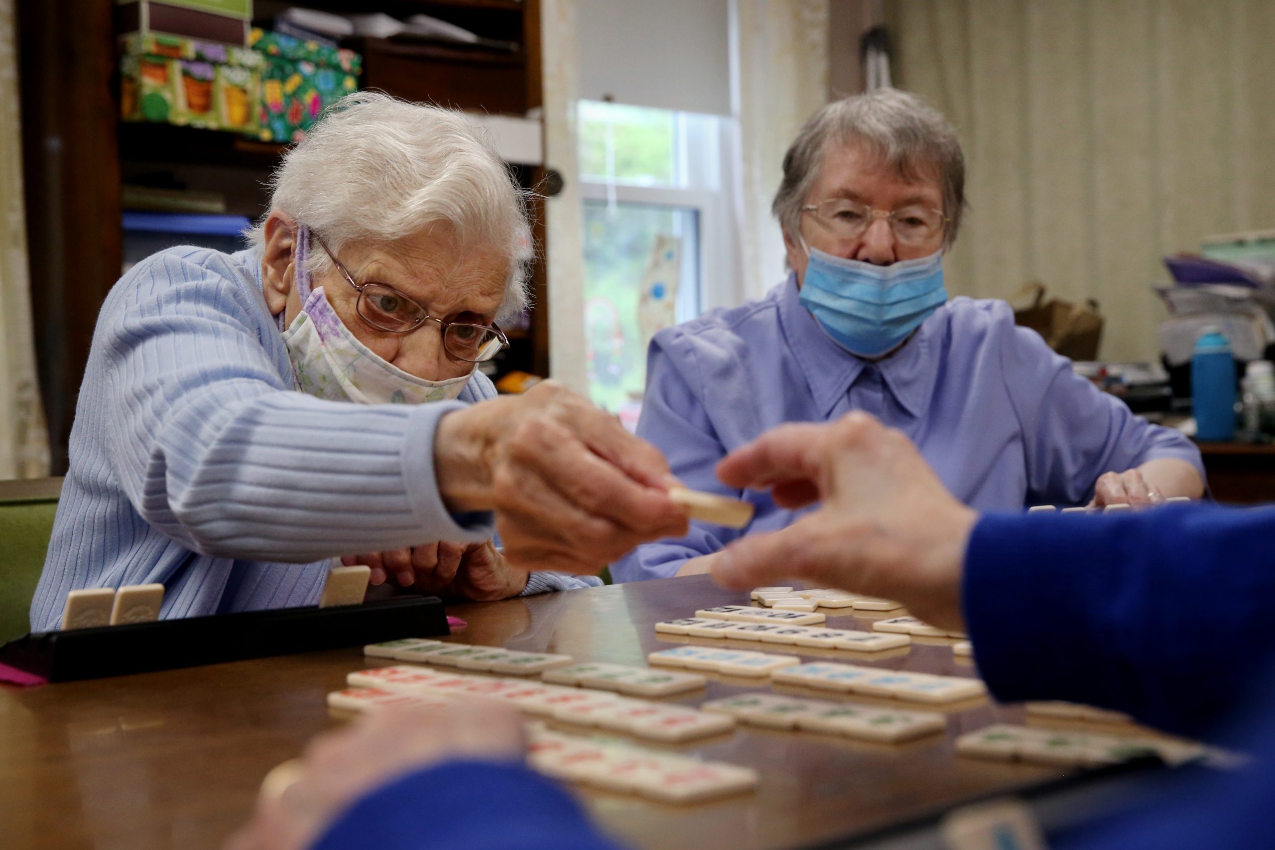 two nursing home residents play a board game at a table. they both wear blue paper face masks.