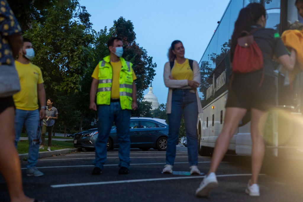 Two men and two women stand outside a bus in D.C. with the Capitol dome in the background. The two men look at the bus door. They wear surgical masks. They wear yellow t-shirts and jeans, one wears a safety vest. Another woman looks on with her arms folded. She wears a yellow t-shirt, a white sweater and jeans. Another woman faces away,looking in the bus door. She wears dark shorts and a dark t-shirt. She wears a red backpack.