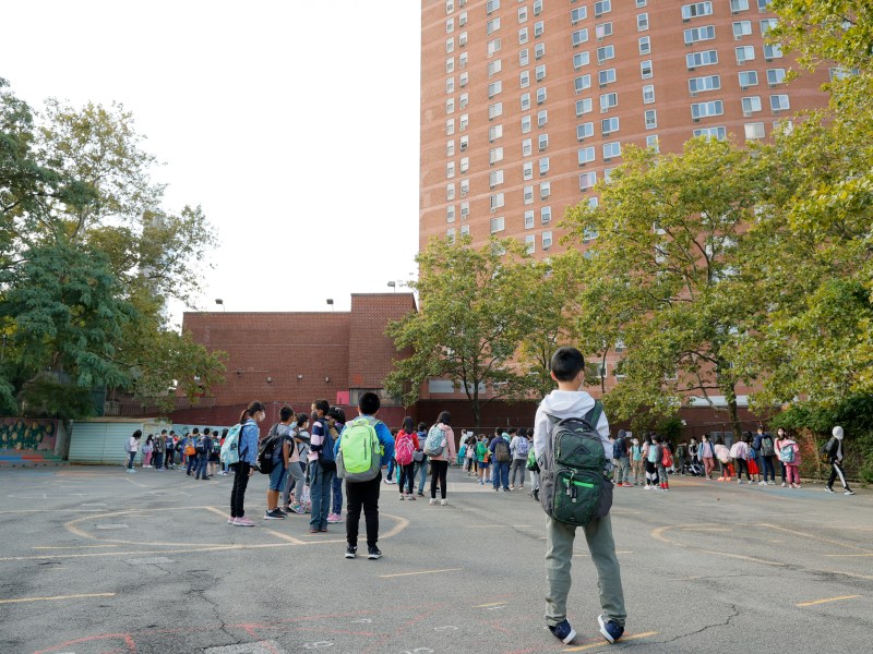 Students stand and walk along the blacktop toward red brick school buildings. There are green trees that line the way.